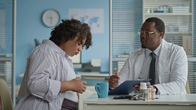 Medium shot of Biracial pregnant woman discussing her concerns with Black male doctor reviewing her chart and medications during prenatal consultation in clinical setting