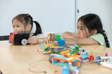 Two Asian girls looking disappointed while working on colorful robot projects, reflecting...