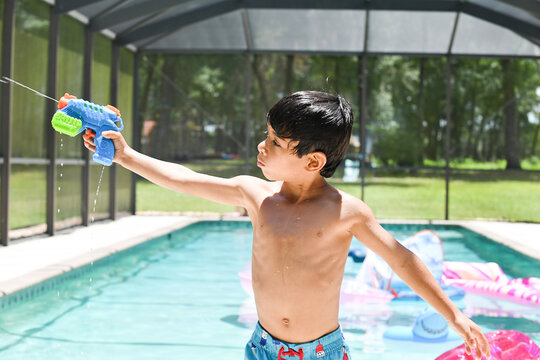 Shirtless boy aims water gun while playing in backyard pool.