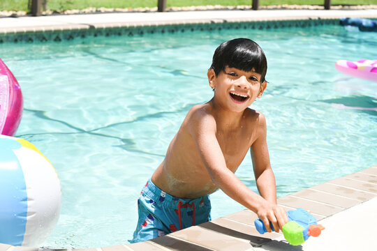 Laughing boy leans on pool edge holding colorful water gun.
