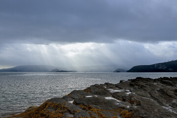 Sunbeams break through clouds over ocean and rocky coastline.