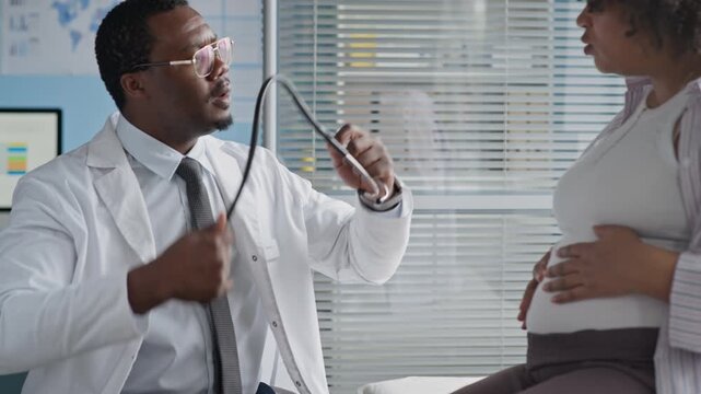 Medium hot of Black male healthcare professional examining pregnant woman using stethoscope in clinical setting, ensuring prenatal care and fetal health monitoring