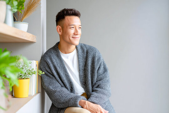 Young man in cozy sweater sitting by window and smiling while enjoying peaceful morning at home