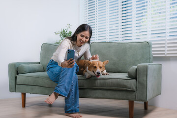 A cheerful Asian woman relaxes on her sofa with her playful corgi, using her smartphone to create...