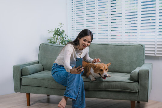 A cheerful Asian woman relaxes on her sofa with her playful corgi, using her smartphone to create engaging social media content, showcasing a cozy lifestyle filled with creativity, joy,companionship