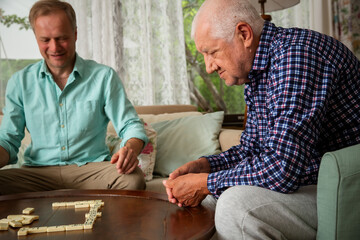 A grown son and his elderly father enjoy a quiet game of dominoes in the living room, sharing connection, laughter, and meaningful time together.