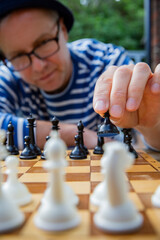 A man in a striped shirt and hat sits in deep thought during an outdoor chess game. Mental challenge under warm summer sun.