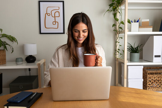 Happy young woman working at home office having coffee. Caucasian female entrepreneur using laptop.