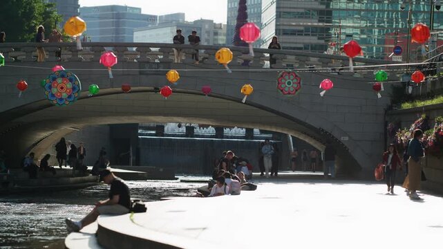 Colorful Lanterns and People Enjoying a Sunny Day by the Water Under a Bridge &mdash; South Korea, Seoul, Cheonggyecheon, downtown, city