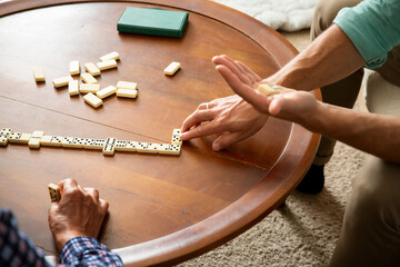 A grown son and his elderly father enjoy a quiet game of dominoes in the living room, sharing connection, laughter, and meaningful time together.