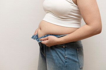 Close-up of a woman in a white tank top struggling to button a pair of tight blue jeans, showing...