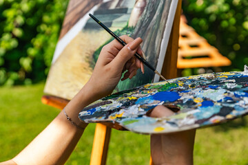 Artist in the middle of painting a picturesque scene. The painting is on an easel, and it includes...