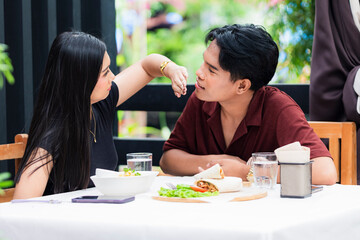 Young Southeast Asian woman playfully blocks her boyfriend’s attempt to kiss her during lunch at...