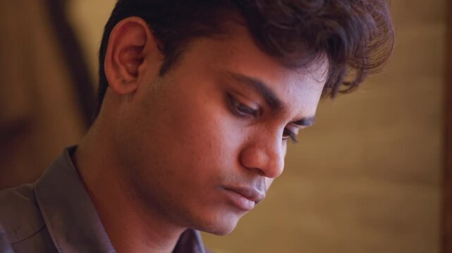 Close-up side profile of young man in natural light looking out window with serious expression, reflecting deep thought or contemplation, captured in quiet indoor setting with soft brown background
