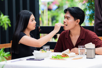 Young girlfriend gently feeds her boyfriend a French fry during their lunch date at an alfresco...