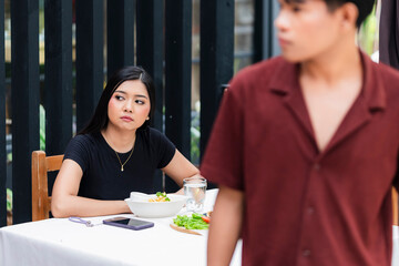 A young and stubborn woman stays behind at a cafe table, watching her boyfriend walk away after a...