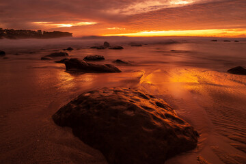 Dramatic golden sunrise casts glowing reflections on wet rocks and sand along a coastal beach.