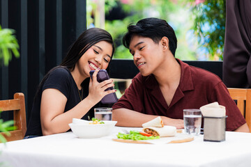 Happy young couple watch funny social media videos on a smartphone during lunch at an alfresco cafe.