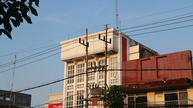 A multi-story building with white facade and windows, next to a shorter building with a red facade, under a blue sky with power lines.