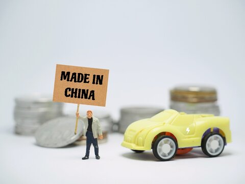 Mini toy of action figure at table with blurred background. Automotive industry photography concept design. Minifigure of people character with Made In China poster banner and Electric Car.