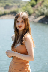 A girl in an orange dress on the shore of a pond, she has long hair falling down her back.