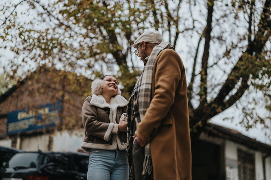 A happy couple enjoys each other's company outside, dressed warmly for a chilly autumn day. Their smiles and laughter are framed by the vibrant fall foliage, creating a heartwarming scene.