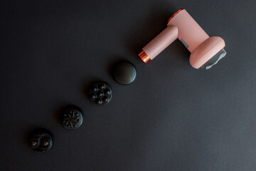 Pink massage gun with soft head and four different black attachments arranged on dark background....