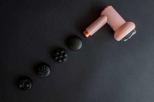 Pink massage gun with soft head and four different black attachments arranged on dark background. Compact muscle recovery device with multiple therapy options