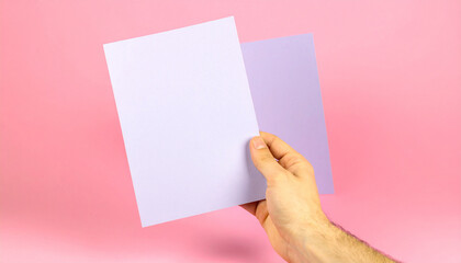 Fototapeta premium Hand Holding Purple Papers on Pink Background: A human hand gracefully presents two sheets of textured purple paper against a soft pink backdrop, creating a study in color, contrast.
