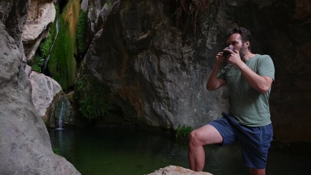 A man finds peaceful contemplation and connection with nature, drinking from a cup by a tranquil waterfall and clear pool in a rocky grotto. This serene scene evokes well-being.