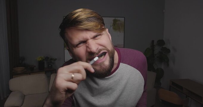 Close-up of a bearded blond man brushing his teeth, looking to the camera, window or tv screen, standing in a room at home with bizarre face, drops his toothbrush