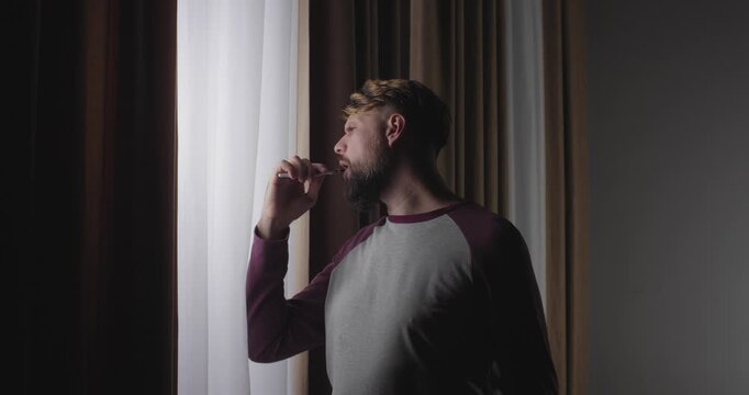 Close-up of a European man, blond and bearded, brushing his teeth in the room looking in the  window. Seeing something  shocking him,  opening his mouth and dropping the brush.