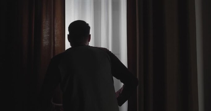 A dark silhouette of a man, seen from behind, standing near a window with curtains, looking out and laughing maliciously at what he sees