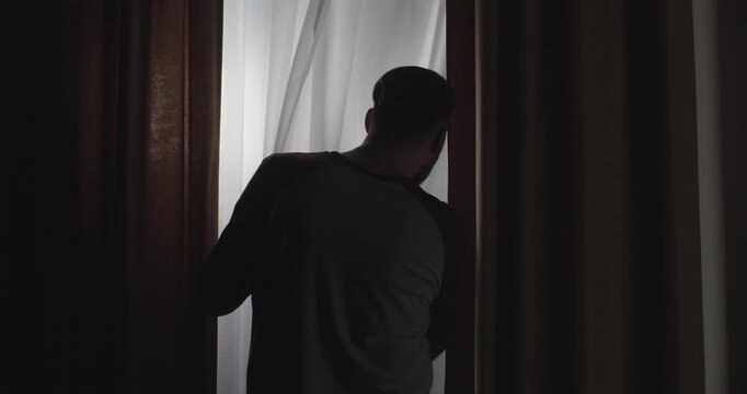 Dark silhouette view from behind of a man walking up to a window, pulling back the curtain and looking out the window trying to see something