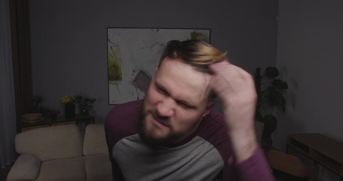 Close-up of a funny, bearded guy looking into the camera, a mirror, or a window, making funny agressive face and fixing his hair with hand