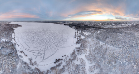 Snow-covered forest on lake shore with ice at sunset and the city on horizon, auerial view