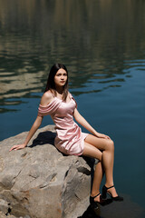 A girl is sitting on the shore of a lake on a large stone, the water in the lake is blue