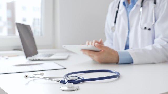 Closeup of blue stethoscope is lying on the table near female doctor reviewing prescription on digital tablet, representing digital healthcare workflow in contemporary medical setting. Medicine