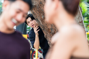 Stunned young man peeks from behind tree, shocked to see ex-girlfriend laughing happily with her...