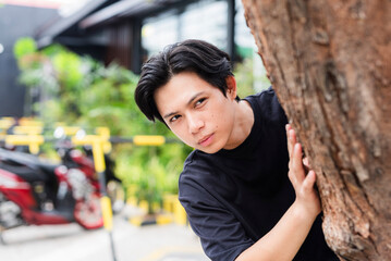 Sneaky young man hiding behind a tree while spying on ex-girlfriend and her new boyfriend during a...