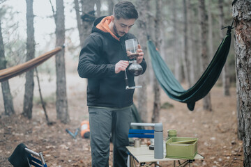 A man brews coffee using a unique setup at a forest campsite. Hammocks hang between trees around...