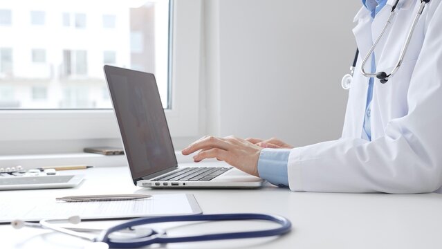 Female doctor is typing on a laptop with a stethoscope, medical record, and pen on the desk in a bright medical office, showcasing healthcare technology. Medicine concept - Powered by Adobe