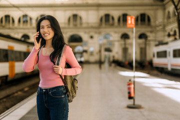 Chinese woman at train station smiling while talking on the phone, surrounded by trains and...