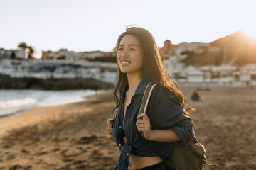 Chinese woman enjoying a sunny day at the beach while carrying a backpack and smiling warmly at the...
