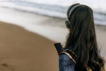 Chinese woman enjoying music while walking along a sandy beach during sunny weather by the ocean