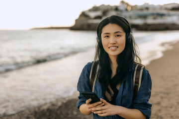 Chinese woman enjoys music while walking along the beach during sunset, capturing a moment of...