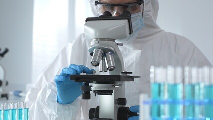A male doctor in a protective suit and gloves uses a microscope while analyzing samples, conducting...