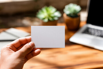 Hand holding blank business card mockup in cozy workspace with notebook. White card template against marble table background. Perfect for branding presentation.