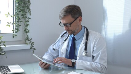 Medical doctor wearing lab coat and stethoscope using digital tablet consulting patient data in...