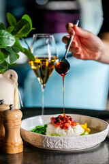 Hand pouring sauce over rice bowl with vegetables and meat. White wine, plant, and wooden shakers in background. Cozy dining scene